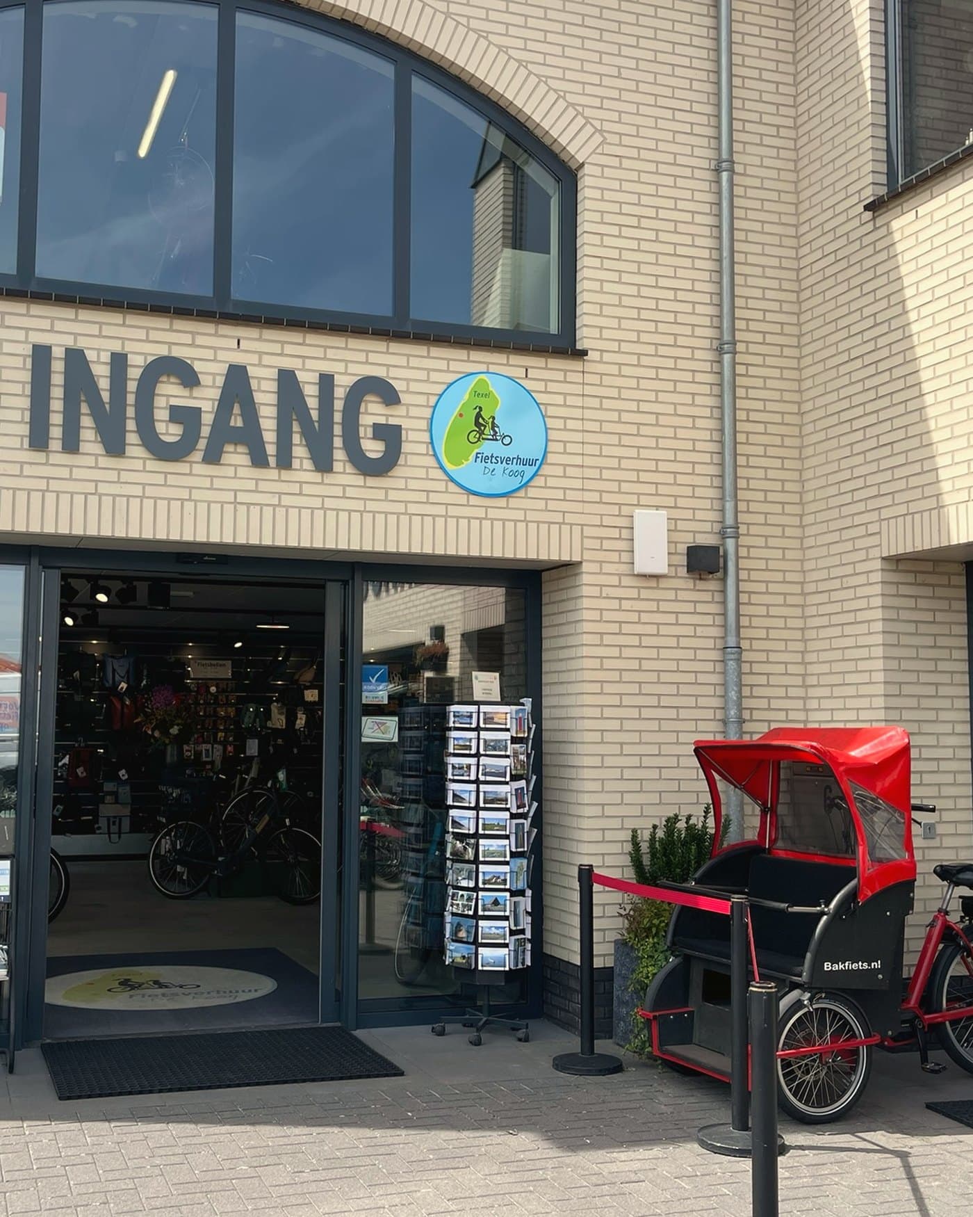 De ingang van Fietsverhuur De Koog met het logo boven de deur en een bakfiets ernaast.