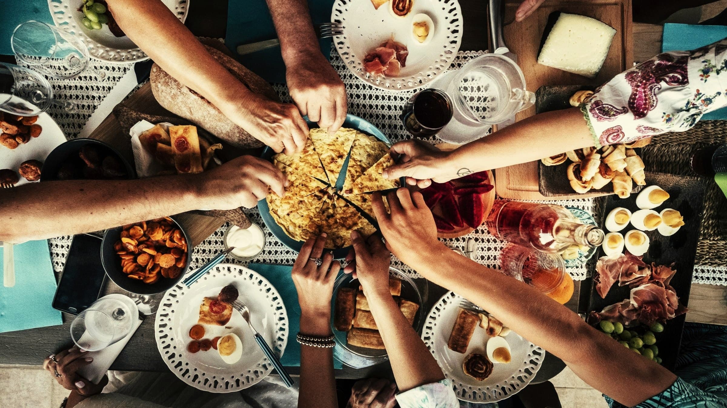 Gedeelde tafel vol gerechten, van bovenaf, handen die een stuk pakken